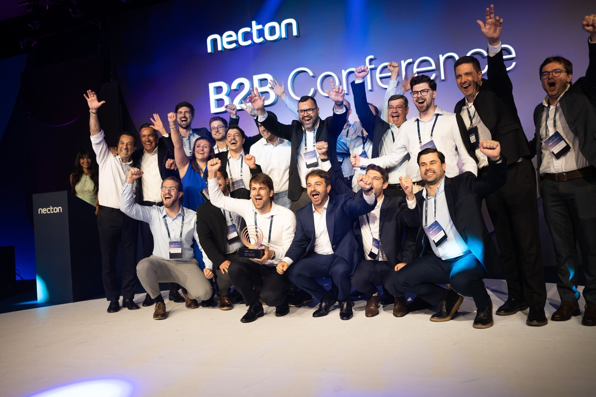 Equipe posando no palco em uma conferência B2B, sorrindo e erguendo os braços em comemoração.